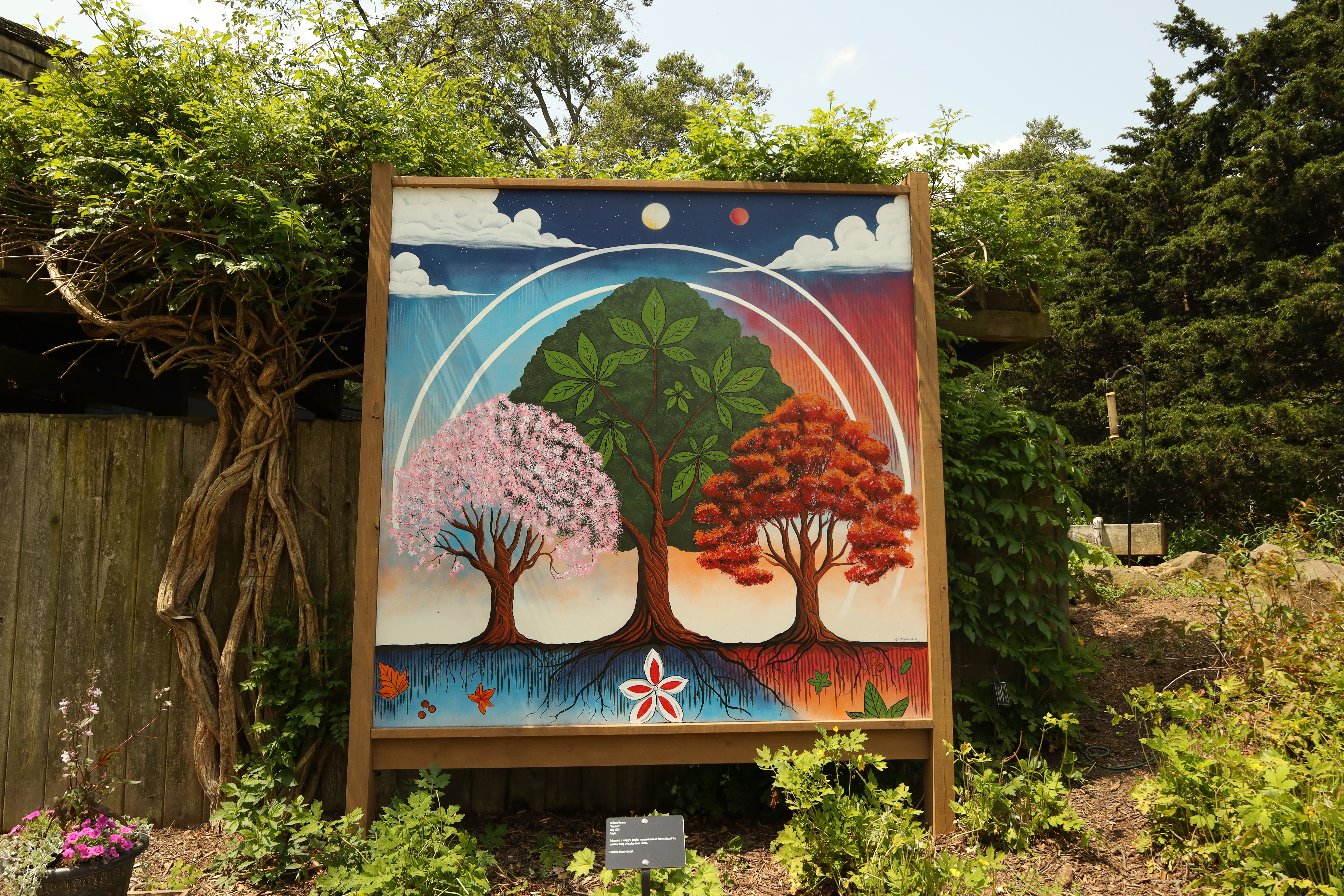 This features three trees. One of the tree tops is pink, one is green and one is orange. it is sitting in The Arboretum surrounded by grass and trees. It has a tan frame.