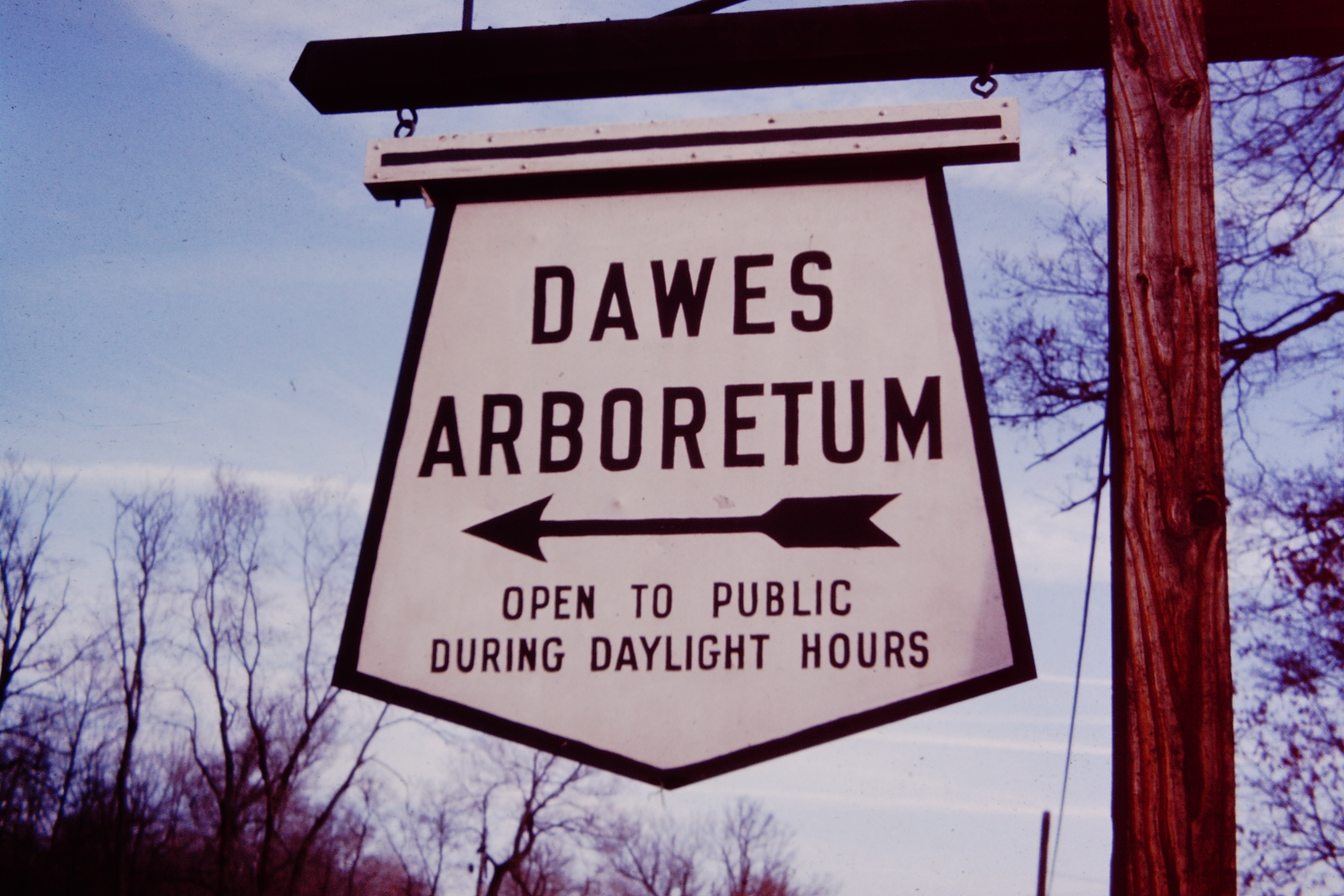 An older sign of The Dawes Arboretum that is black and white with an arrow pointing to the light. It reads "Dawes Arboretum: open to public during daylight hours." It's hung on a brown post.
