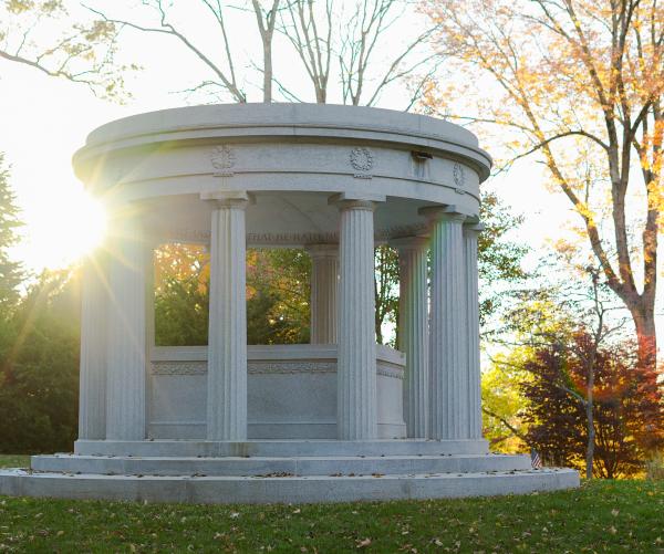 A white memorial with four visible pillars stands with the sunset behind it. The trees are green on the left and orange on the right.