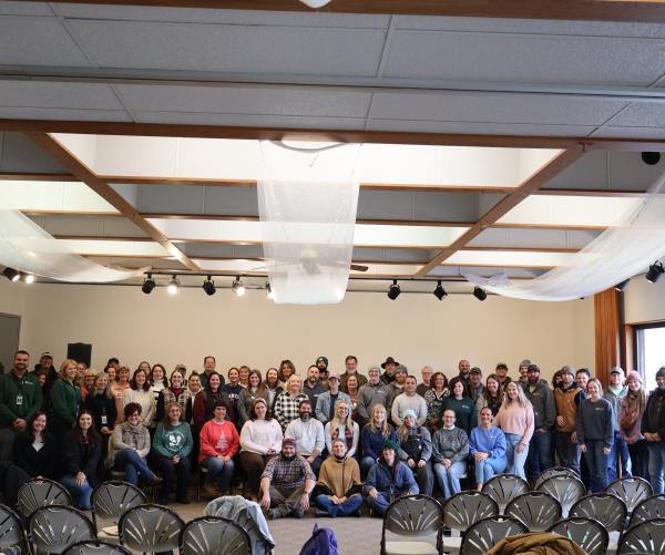 A large group of people, about 80 individuals, stand against a tan wall. The cieling is tan with white flowy curtains. There is a window with natural light shining through on the right side of the photo.