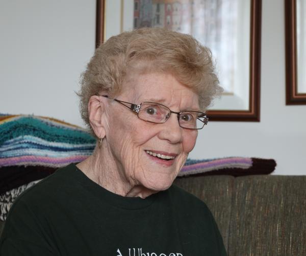 A light complexion female with short blonde hair and square glasses smiles at the camera and is wearing a dark green shirt.