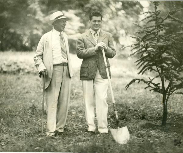 Beman Dawes, co-founder of The Arboretum and a Tree Dedicator stand together with a shovel near a tree. They are on top of grass and surrounded by trees. This is a black and white photo. 