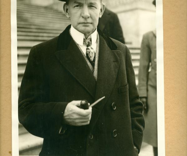 Charles Dawes stands in a black-and-white in a suit, with a tie, a tophat and he's holding a cigar. He's standing in front of a large collection of stairs.