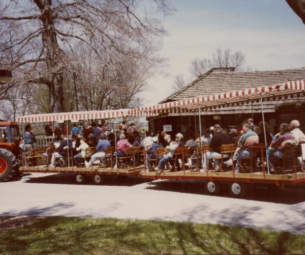 A group of people sit on a wagon being pulled by an orange tractor. The tractors have striped roofs and the wagons are sitting in front of the Visitors Center.