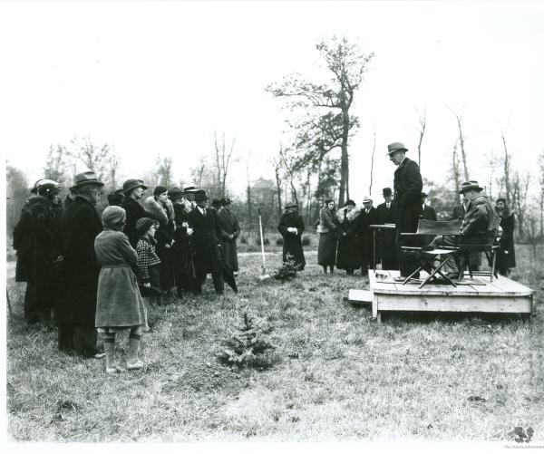 A black and white photo showcases a group of over 10 people watching a man stand on a wooden platform and talking in front of a tree. They are all among other trees, dressed in warm winter clothes.