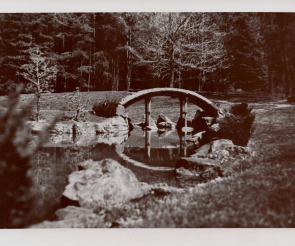 This is a black and white photo of The Dawes Arboretum. It shows a bridge over water with many trees surrounding it.