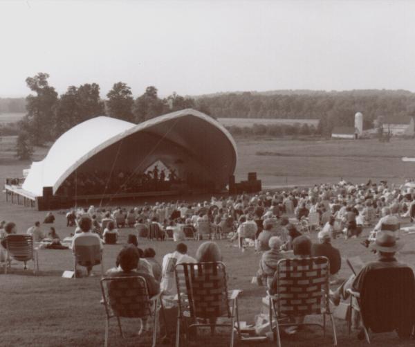 A gray and white photo shows a large white amphiteahter and groups of people gathered sitting on chairs listening in front of it.