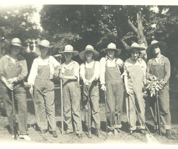 This is a black and white photograph of a group of women wearing white shirts, overalls and sun hats while holding shovels outside.