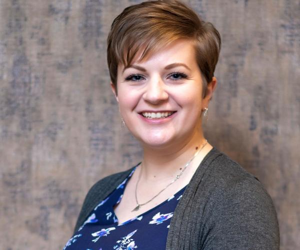 Holly Latteman stands proudly with a blue shirt with a design on it with a grey sweater and a silver necklace. The background is grey. She is fairskinned with blonde hair. 