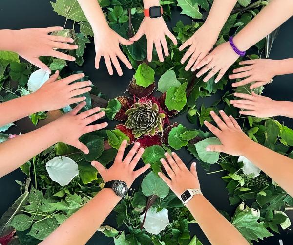 A group of hands surround greenery at The Dawes Arboretum at Camp Dawes. There are twelve hands surrounding the greenery with something purple in the middle.
