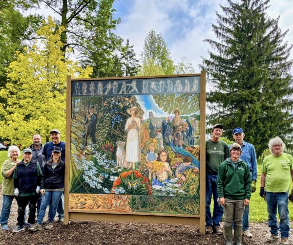 Seven Dawes volunteer and employees stand on both sides of a mural for The Dawes Arboretum's Year of Art. They are surrounded by green trees and are wearing green shirts.