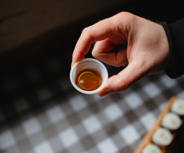 A hand holds a small, white plastic cup of maple syrup that's a bit larger than a quarter. There is plaid brown and white tablecloth underneath it that is out of focus. The hand and maple syrup cup our in focus. Whomever is holding the cup has a black sleeve. 