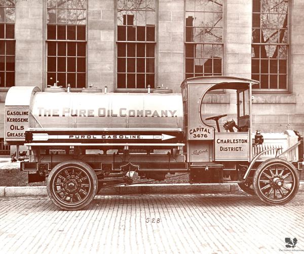 A black and white photo of a Pure Oil truck that says "The Pure Oil Company" sits on a brick road in front of five large vertical windows.