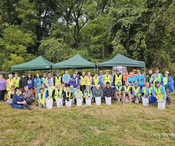 Approximately 50 volunteers are sitting and standing on a grassy area with three green tents behind them. Half of them are wearing bright yellow safety vests, and a lot of them are wearing blue t-shirts. There are green trees above them. They are smiling.