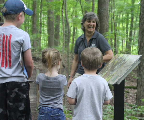 A tan woman stands admist green trees and leans against a stantion with information. She has grey hair and is wearing a grey shirt. Three kids stand in front of her listening intently, their backs are to the camera. A man with a brown hat stands in the background and smiles, listening to the women and watching the kids.