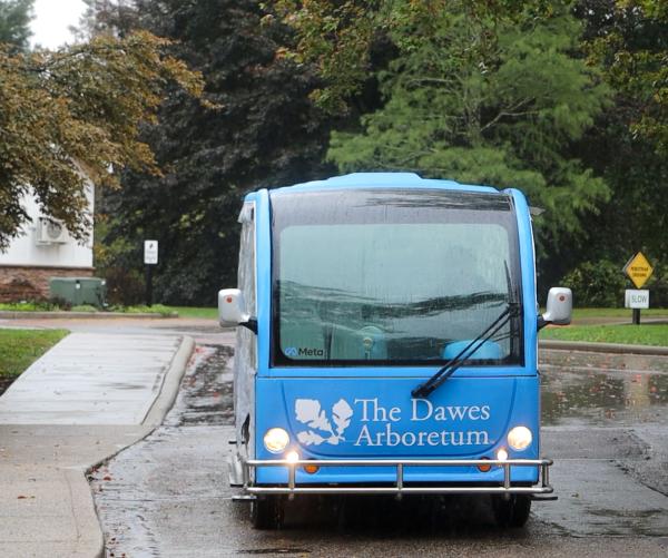 A large blue electric tram sits idle int the rain surrounded by green trees. It is blue with a white Dawes Arboretum logo on the front of it.