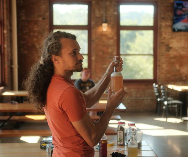 A caucasian man wearing a salmon shirt stands and holds a large, glass bottle of fermented soda.