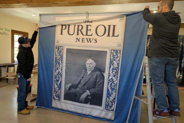 Two people wearing black shirts and blue pants each stand on a side of a large, blue, white and black fabric printing of Beman Dawes in front of The Pure Oil News.