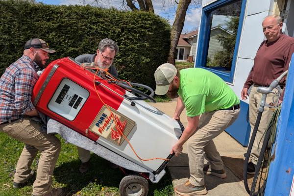 A group of Dawes employees carry a red and white Pure Oil gas station pump while a man stands behind them and looks at the camera.