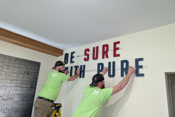 Two Dawes employees, both wearing neon green shirts, hang up a Be Sure with Pure sign in The Dawes Arboretum's History Center.