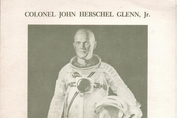 This is a black-and white photo of John Glenn, an astronaut. He's wearing a space suit and holding a helmet.