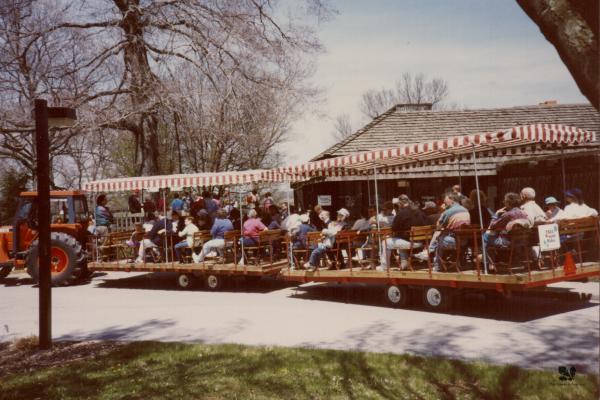 Groups of people sit in a wagon with a range and white top in front of The Arboretum's Visitors Center.