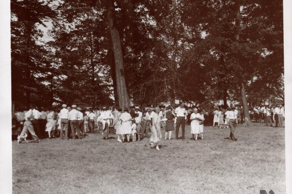 This is a black-and-white photo of many people on The Arboretum's grounds during an annual picnic hosted at Dawes. They are surrounded by trees, are standing on grass and are dressed in nice clothing.