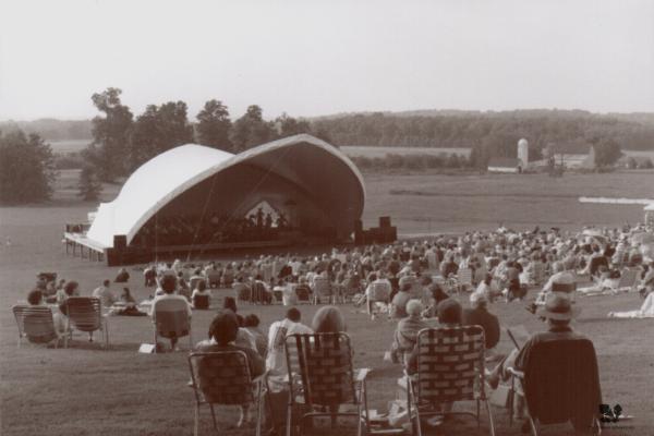 Large groups of people sit in front of a large white canopy, an auditorium, outside on The Arboretum's grounds. People are sitting on chairs and in blankets listening to music. Trees dot the background.