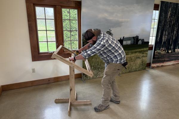 A Dawes employee wearing a black hat and a plaid blue and white shirt measures a wooden frame.