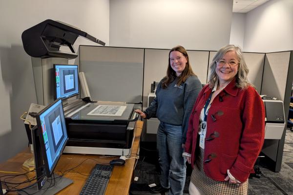 Two fair-skinned women smile at the camera in front of a large scanner at the Columbus Metropolitan Library.