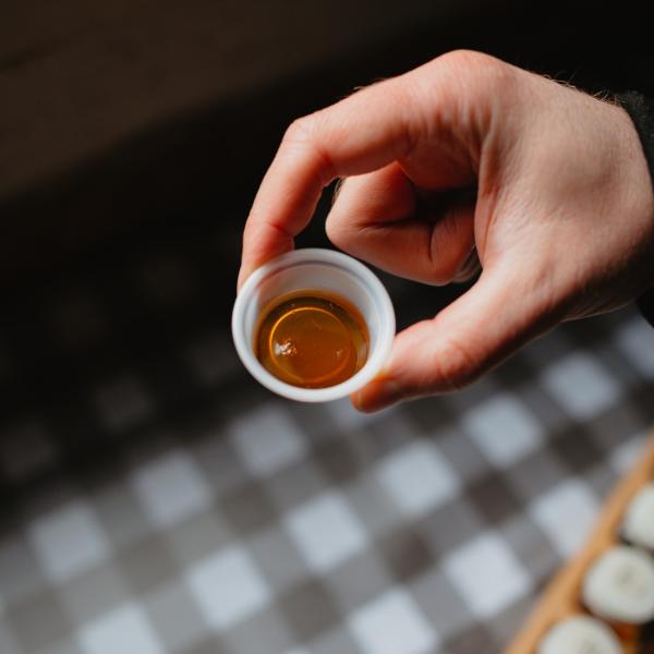 A hand holds a small, white plastic cup of maple syrup that's a bit larger than a quarter. There is plaid brown and white tablecloth underneath it that is out of focus. The hand and maple syrup cup our in focus. Whomever is holding the cup has a black sleeve. 