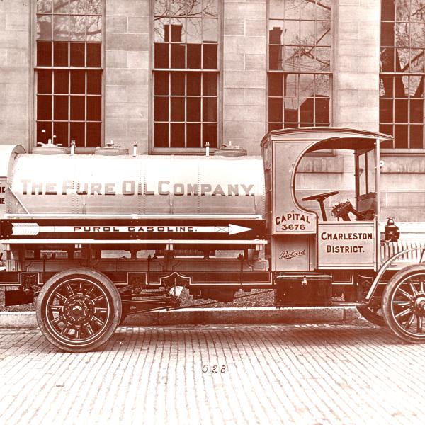 A black and white photo of a Pure Oil truck that says "The Pure Oil Company" sits on a brick road in front of five large vertical windows.