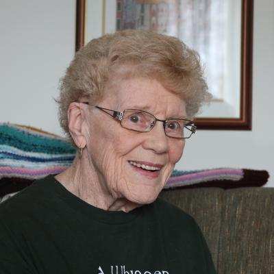 A light complexion female with short blonde hair and square glasses smiles at the camera and is wearing a dark green shirt.