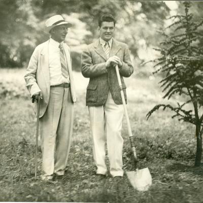 Beman Dawes, co-founder of The Arboretum and a Tree Dedicator stand together with a shovel near a tree. They are on top of grass and surrounded by trees. This is a black and white photo. 