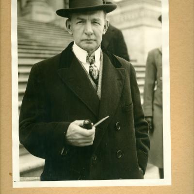 Charles Dawes stands in a black-and-white in a suit, with a tie, a tophat and he's holding a cigar. He's standing in front of a large collection of stairs.