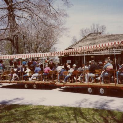 A group of people sit on a wagon being pulled by an orange tractor. The tractors have striped roofs and the wagons are sitting in front of the Visitors Center.