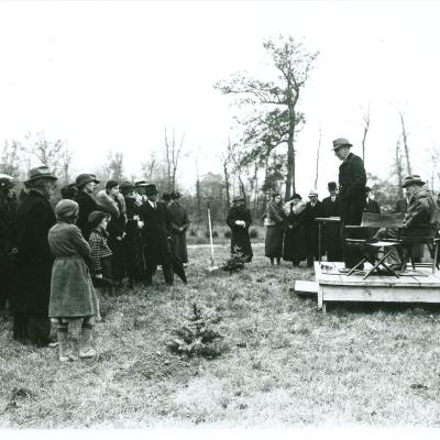 A black and white photo showcases a group of over 10 people watching a man stand on a wooden platform and talking in front of a tree. They are all among other trees, dressed in warm winter clothes.