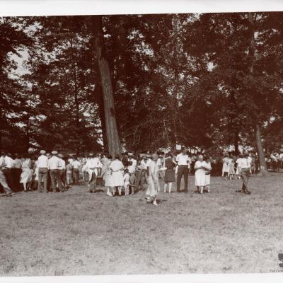 This is a historical photo of The Pure Oil Company's annual picnic. It is black and white, and shows over 100 people walking around the grounds of the farm. Surrounded by tall trees, the people are in professional clothing and talking amongst one another.