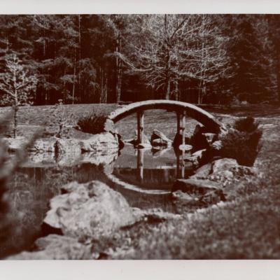 This is a black and white photo of The Dawes Arboretum. It shows a bridge over water with many trees surrounding it.