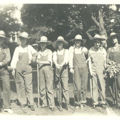 This is a black and white photograph of a group of women wearing white shirts, overalls and sun hats while holding shovels outside.