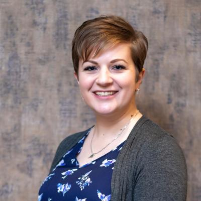 Holly Latteman stands proudly with a blue shirt with a design on it with a grey sweater and a silver necklace. The background is grey. She is fairskinned with blonde hair. 
