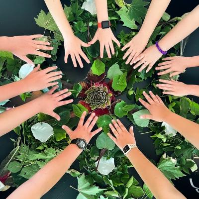 A group of hands surround greenery at The Dawes Arboretum at Camp Dawes. There are twelve hands surrounding the greenery with something purple in the middle.