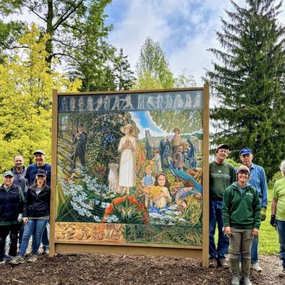 Seven Dawes volunteer and employees stand on both sides of a mural for The Dawes Arboretum's Year of Art. They are surrounded by green trees and are wearing green shirts.