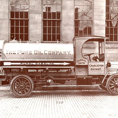 A black and white photo of a Pure Oil truck that says "The Pure Oil Company" sits on a brick road in front of five large vertical windows.