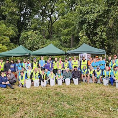 Approximately 50 volunteers are sitting and standing on a grassy area with three green tents behind them. Half of them are wearing bright yellow safety vests, and a lot of them are wearing blue t-shirts. There are green trees above them. They are smiling.