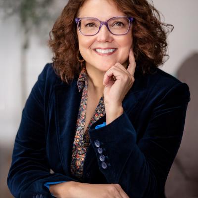 Stephanie Crockatt, Executive Director of The Dawes Arboretum, sits on a chair wearing a navy blue balzer. She is wearing glasses with her left hand on her chin.
