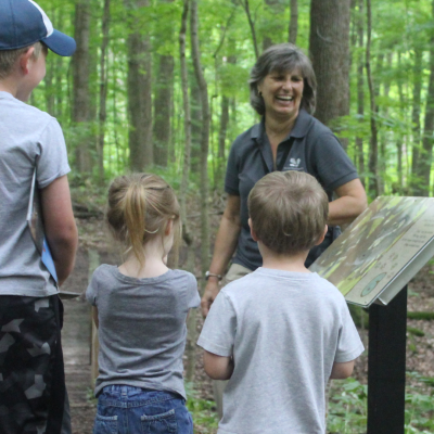 A tan woman stands admist green trees and leans against a stantion with information. She has grey hair and is wearing a grey shirt. Three kids stand in front of her listening intently, their backs are to the camera. A man with a brown hat stands in the background and smiles, listening to the women and watching the kids.