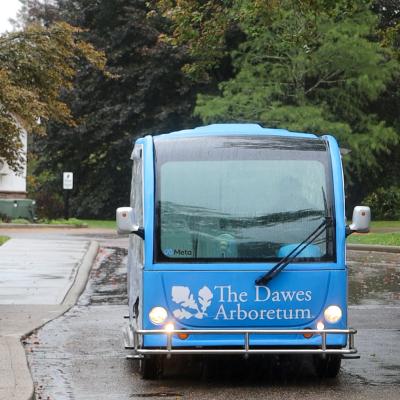 A large blue electric tram sits idle int the rain surrounded by green trees. It is blue with a white Dawes Arboretum logo on the front of it.