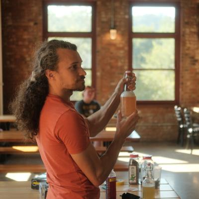 A caucasian man wearing a salmon shirt stands and holds a large, glass bottle of fermented soda.