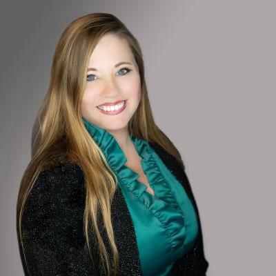 A light complexion female with blue eyes and blondish brown hair wearing a dark green blouse with ruffles and a black cardigan smiles at the viewer.